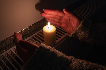 A burning candle with women's hands frozen from frost on the radiator heating in dark home. Shutdown of heating and electricity, power outage, blackout, load shedding or energy crisis, concept image.