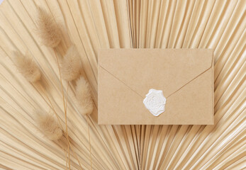Blank sealed envelope on dry beige palm leaf near hare's tail grass top view,  boho wedding mockup