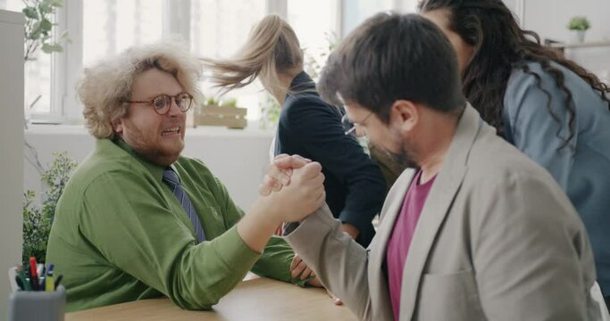 Funny chubby guy is arm wrestling with coworker in office winning having fun in workplace together. Corporate activities and youth lifestyle concept.