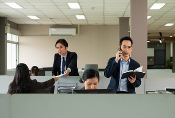 Businessman in dark blue suit with eyeglasses making a call on mobile phone and checking information or organizing event on  notebook while standing in open space office with blurred colleagues workin