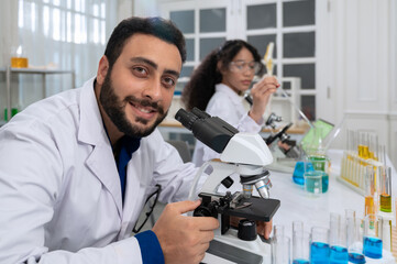 Smiling male scientist with beard in white laboratory coat working with microscope for research in medical development laboratory .