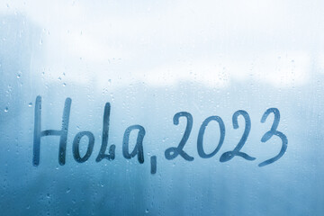 lettering Hola in Spanish is hello in english and numbers 2023 paint with finger with streaks of water on splashed by rain foggy glass on blue window © ksenija1803z