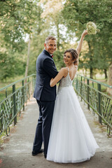 Glad smiling wedding couple standing on bridge in park in summer, looking back. Young bride...