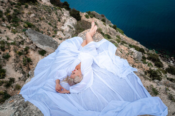 The blonde lies on a rock above the sea. With long hair on a sunny seashore in a white dress, rear view, silk fabric flutters in the wind. Against the backdrop of blue skies and mountains.