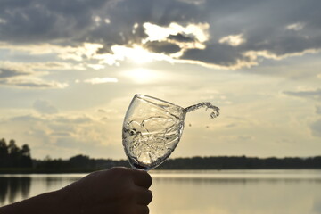 A man's hand holds a glass from which water flows against the backdrop of a sunset on the lake.