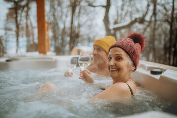 Senior couple in kintted cap enjoying together outdoor bathtub and enjoying glass of wine at their terrace during cold winter day.