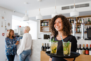Happy young waitress with tray serving cocktail drinks at restaurant