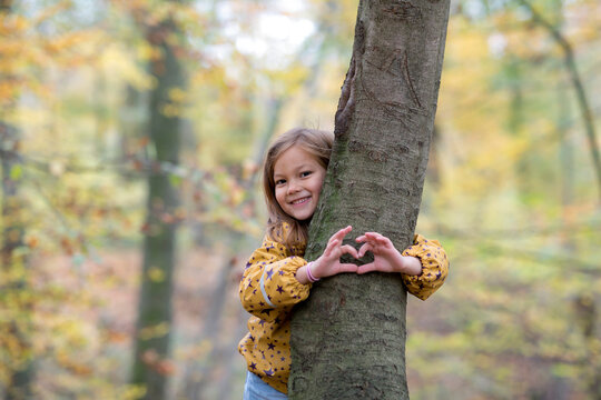 Smiling cute girl hugging tree and gesturing heart shape in forest