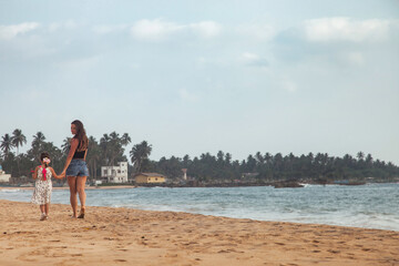 Rear view mom and little daughter together walking on tropical sandy beach at sea background. Mother and little girl in casual clothes on ocean. Concept family vacation, travel. Copy text space
