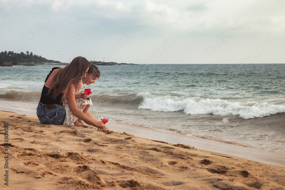 Wall mural rear view mom and little daughter together sitting on tropical sandy beach at sea background. mother