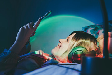 Happy woman wearing wireless headphones using smart phone lying on bed at home