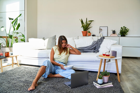 Woman holding bowl looking at laptop by sofa in living room