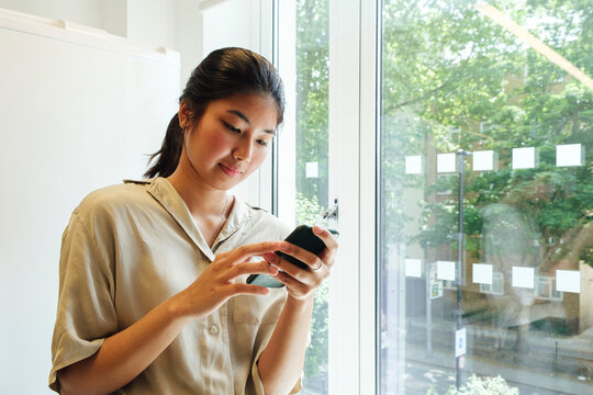 Xoung Woman Standing At The Window Reading Text Messages On Her Smartphone