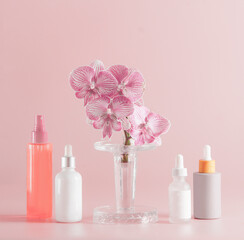 Modern cosmetic products setting with various bottles and orchid flowers at pastel pink background. Front view. Beauty concept