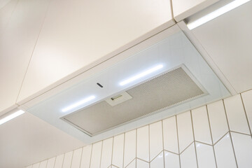 A narrow white glossy hood over the hob with backlight against the background of white tiles and...