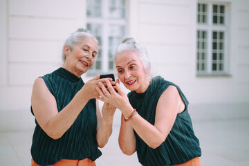 Senior women twins outdoors in city checking smartphone.