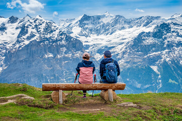 Unknown couple sits on wooden bench and enjoys the view of Swiss Alps at Hiking trail from First peak to Bachalpsee. Grindelwald.