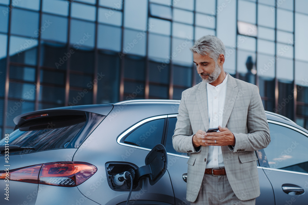 Wall mural businessman holding smartphone while charging car at electric vehicle charging station, closeup.