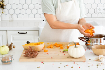Woman hands putting a butternut diced carrot into the saucepan in modern white kitchen. Homemade autumn pumpkin soup recipe