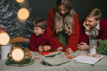 Young parents with their cute boy preparing Christmas cookies in the kitchen. Favorite family tradition. Christmas lights on foreground.