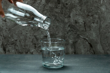 Detail of the hand of an unrecognizable man pouring water from a glass bottle into a glass on a textured background.