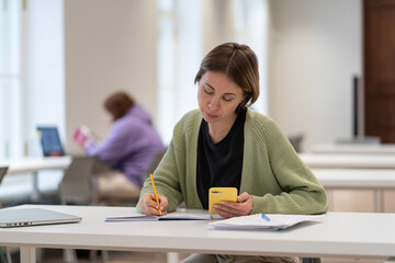 Obraz na płótnie Canvas Concentrated middle-aged scandinavian female student using smartphone for exam preparation while sitting at library table. Focused woman mature student studying, learning online, taking second degree