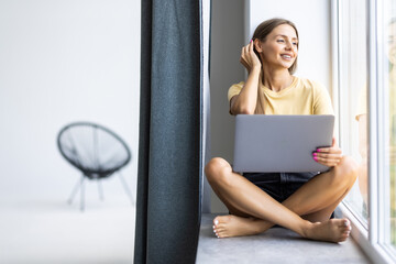 Young pretty woman sitting on the windowsill and working online using her laptop in the room