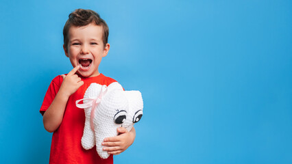 A smiling boy with healthy teeth holds a plush tooth in his hands on a blue isolated background....