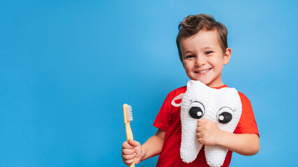 A smiling boy with healthy teeth holds a plush tooth and a toothbrush on a blue isolated...