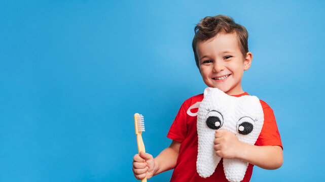 A smiling boy with healthy teeth holds a plush tooth and a toothbrush on a blue isolated background. Oral hygiene. Pediatric dentistry. Prevention of caries. A place for your text.