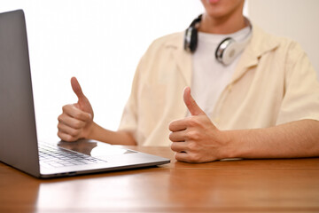 Asian male looks at his laptop screen and makes a thumbs up hand gesture. cropped image