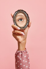 Retro style. Female hand holding small mirror with reflection of girl's eye isolated over pink background. Concept of style, beauty, art