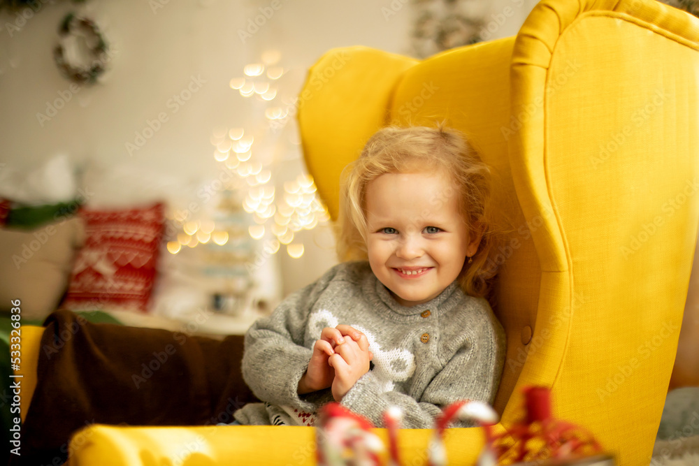 Wall mural Cute little toddler curly girl in winter christmas sweater, sitting in cozy room, decorated for christmas