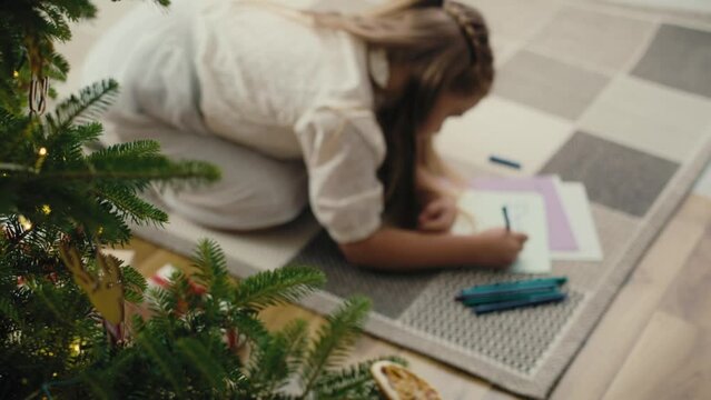 Top view of little caucasian girl lying on carpet next to Christmas tree and writing a letter to Santa Claus. Shot with RED helium camera in 8K.   