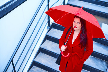 Young executive business woman covers herself with a red umbrella to protect herself from the rain on a cloudy winter day as she walks down the stairs. Blue Monday concept.