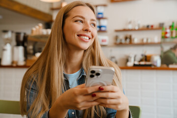 White blonde woman smiling and using mobile phone while sitting in cafe