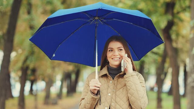 Beautiful young woman with blue umbrella talking on the phone while walking through the rainy autumn park. Blurred trees on background. Concept of leisure