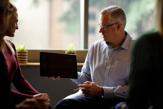 Mature caucasian businessman presenting information on digital tablet to team sitting in a meeting in office. Mature manager leading team of coworkers.