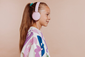 Side view of cute little kid girl 10 years old listen music with headphones, isolated on pastel beige background. Childhood lifestyle concept. Mock up copy space.