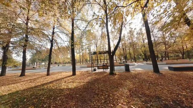 FPV drone view of A guy and a girl swing on a swing in the autumn park. People are walking. The leaves on the trees are orange, yellow and red. There are benches, flower beds, lanterns in the park. Th