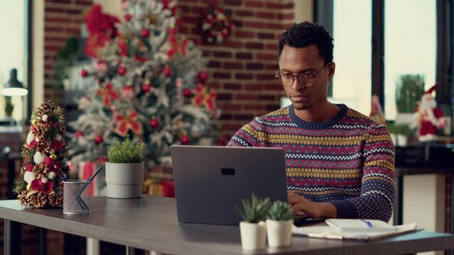Company employee using laptop in christmas decorated office filled with tree and lights for winter festivity. Young adult working with documents at desk during xmas holiday season.