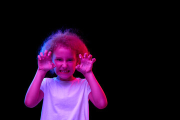 Child is angry. Little girl, kid in white t-shirt isolated over dark background in pink neon light. Concept of children emotions