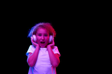 Little shocked girl 6-7 years old wearing white t-shirt and headphones listen to music isolated on dark background in neon light.