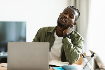 Tired black man suffering from neck spasm while working on laptop and rubbing his muscles, sitting at desk