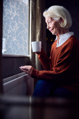 Senior Woman With Hot Drink Trying To Keep Warm By Radiator At Home In Cost Of Living Energy Crisis