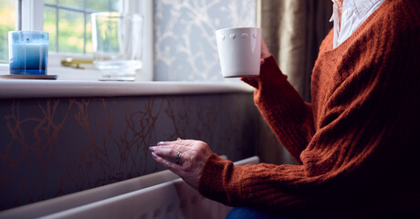 Senior Woman With Hot Drink Trying To Keep Warm By Radiator At Home In Cost Of Living Energy Crisis