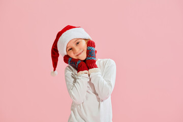 Emotional charming girl, child in casual style clothes, knitted mittens and Christmas hat having fun over pink background. Christmas, New Year, dreams, holidays