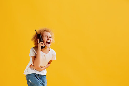 Laughing. Cute little girl, kid using retro mobile phone isolated over yellow background. Concept of children emotions, fashion