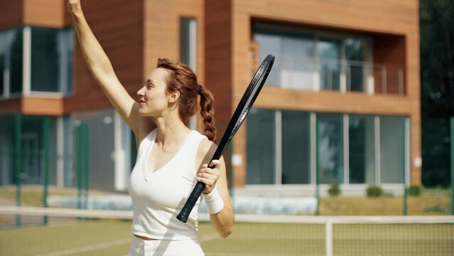 Professional female tennis player thanks match visitors. Woman in white on grass court with racket celebrating win. Sunny day tennis club concept.