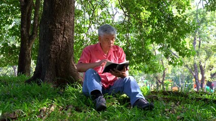 Happy Asian old senior man with gray hair reading book outside in park. Concept of happy elderly man after retirement and good health